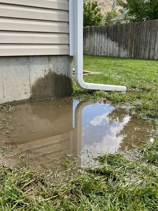 Rainwater pooled against a home’s concrete foundation below a downspout that discharges at the wall