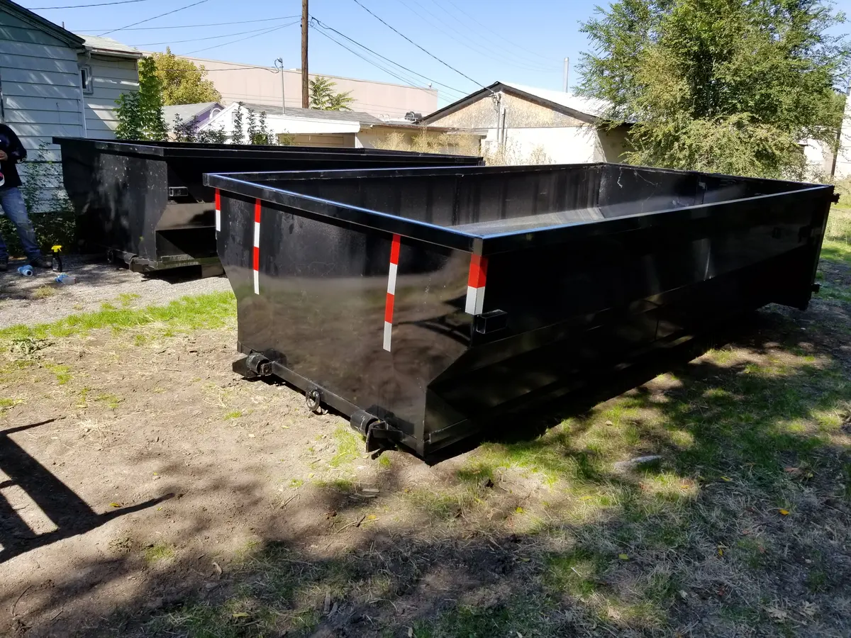 Two black open-top roll-off dumpsters with red and white reflective striping, dropped side by side on a residential lawn