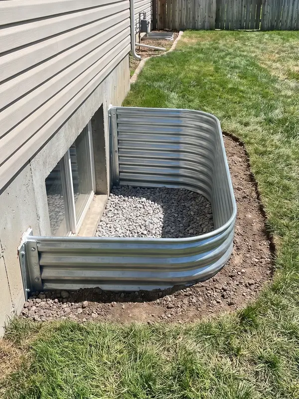 Newly installed galvanized window well bolted and sealed against a home’s concrete foundation, its base filled with washed drainage gravel