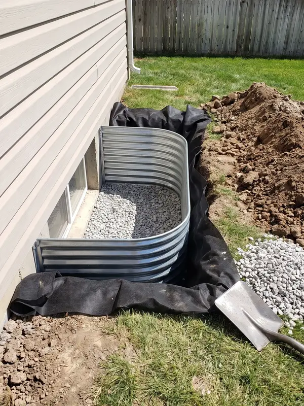 Window well installation in progress with filter fabric lining the excavation and washed drainage gravel going into the base around a basement window