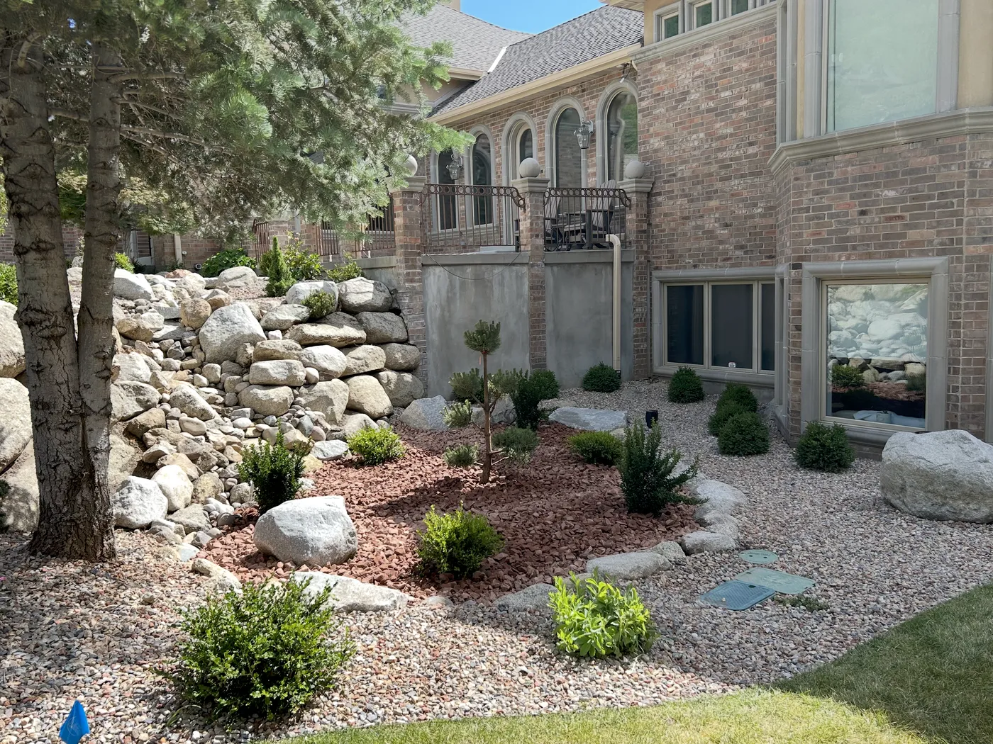 Boulder retaining wall above a fresh red-stone bed with shaped shrubs and young pines, the brick home behind