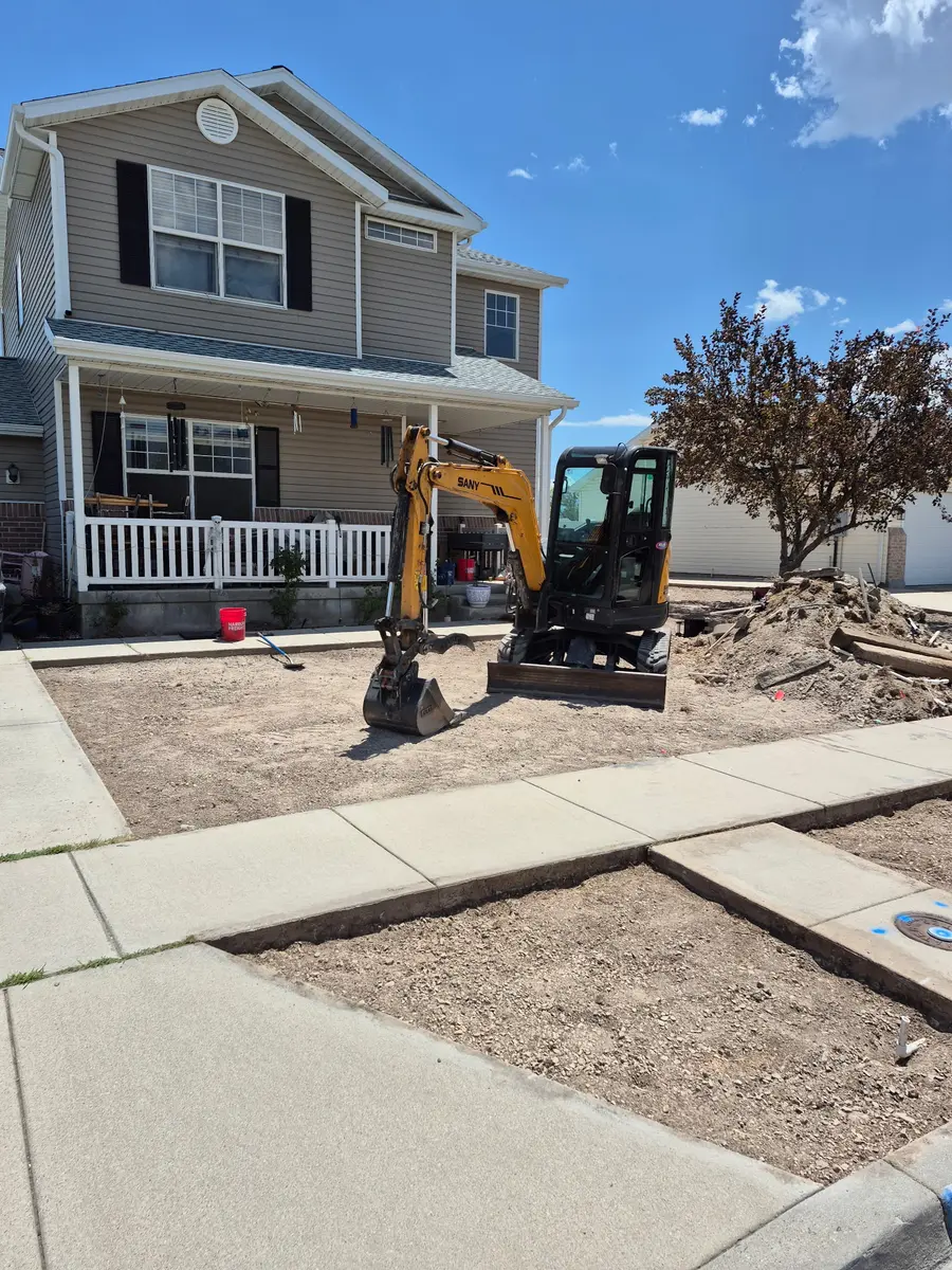 The front yard stripped back to bare dirt with a mini-excavator parked mid-dig and the two-story home behind