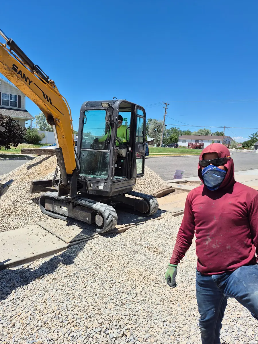 A mini-excavator parked on construction mats and plywood laid over the walkway, with a crew member crossing the fresh drainage rock