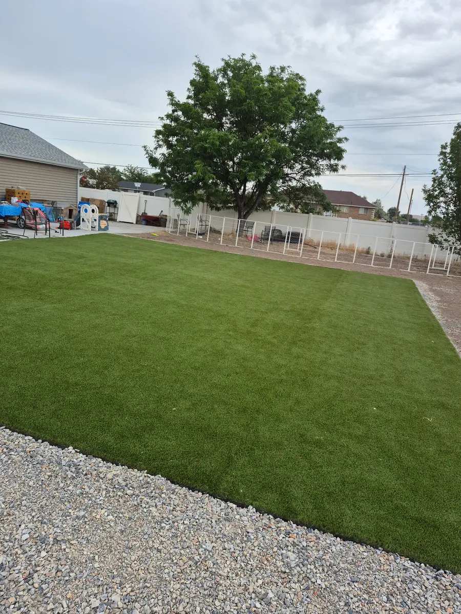 Finished backyard with artificial turf laid wall to wall inside a white vinyl fence, a mature shade tree at the back and a crushed-rock border in front