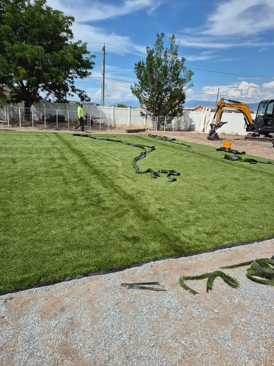 Artificial turf rolled out across the backyard mid-install with the seams still open and a mini-excavator working at the fence line